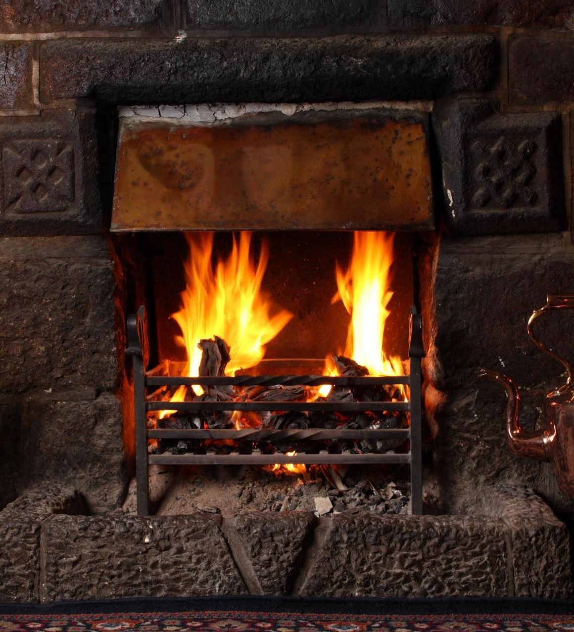 A cozy open fireplace with a roaring fire set in a rustic stone hearth. A copper kettle sits nearby, reflecting the warm glow, alongside traditional fireplace tools.