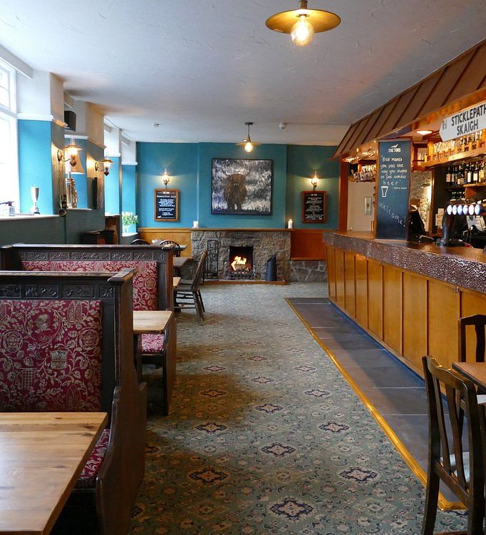 A cozy traditional pub with wooden booths, a well-stocked bar, and a lit fireplace. The interior features teal walls, warm lighting, and a welcoming atmosphere.