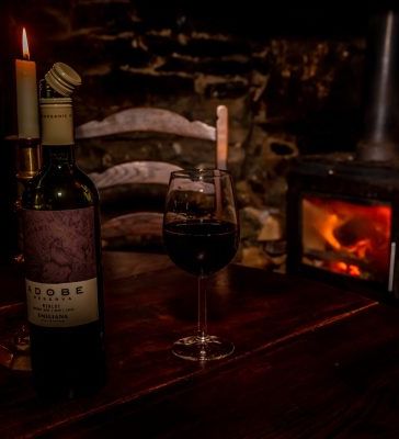 A dimly lit rustic setting with a bottle of red wine, a glass, and a burning candle on a wooden table. A wood-burning stove in the background adds warmth and ambiance.