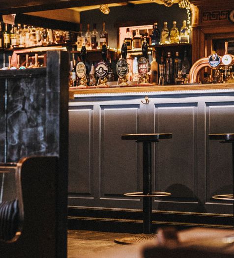 A cozy pub interior with rustic wooden booths, patterned cushions, and a well-stocked bar featuring beer taps and spirits. A sign above the bar reads 