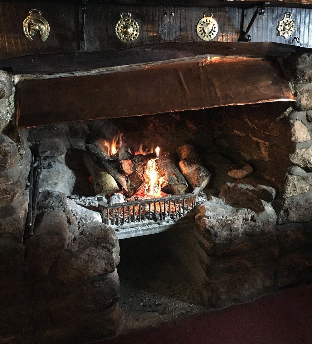 A rustic open fireplace with a crackling log fire, set in a stone hearth with a copper hood. Above, chalkboard menus and brass decorations add to the cozy pub ambiance.