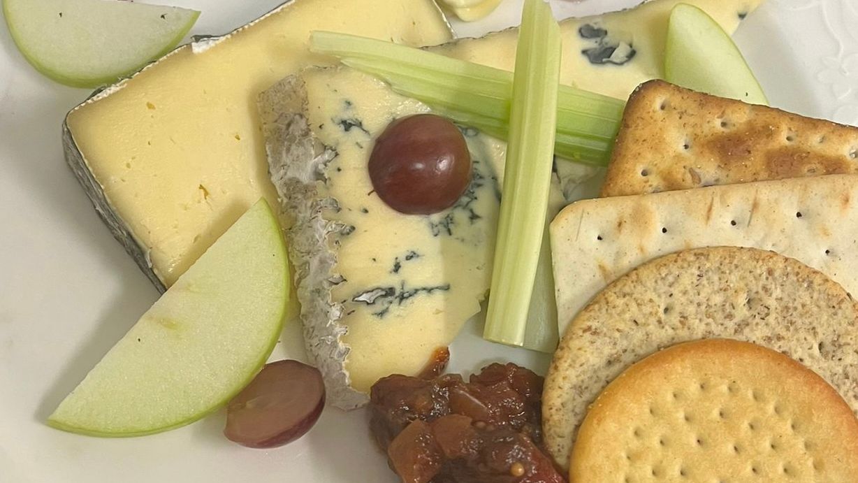 Cheese platter with crackers, apple slices, grapes, celery, chutney, and a swirl of butter on a white plate.