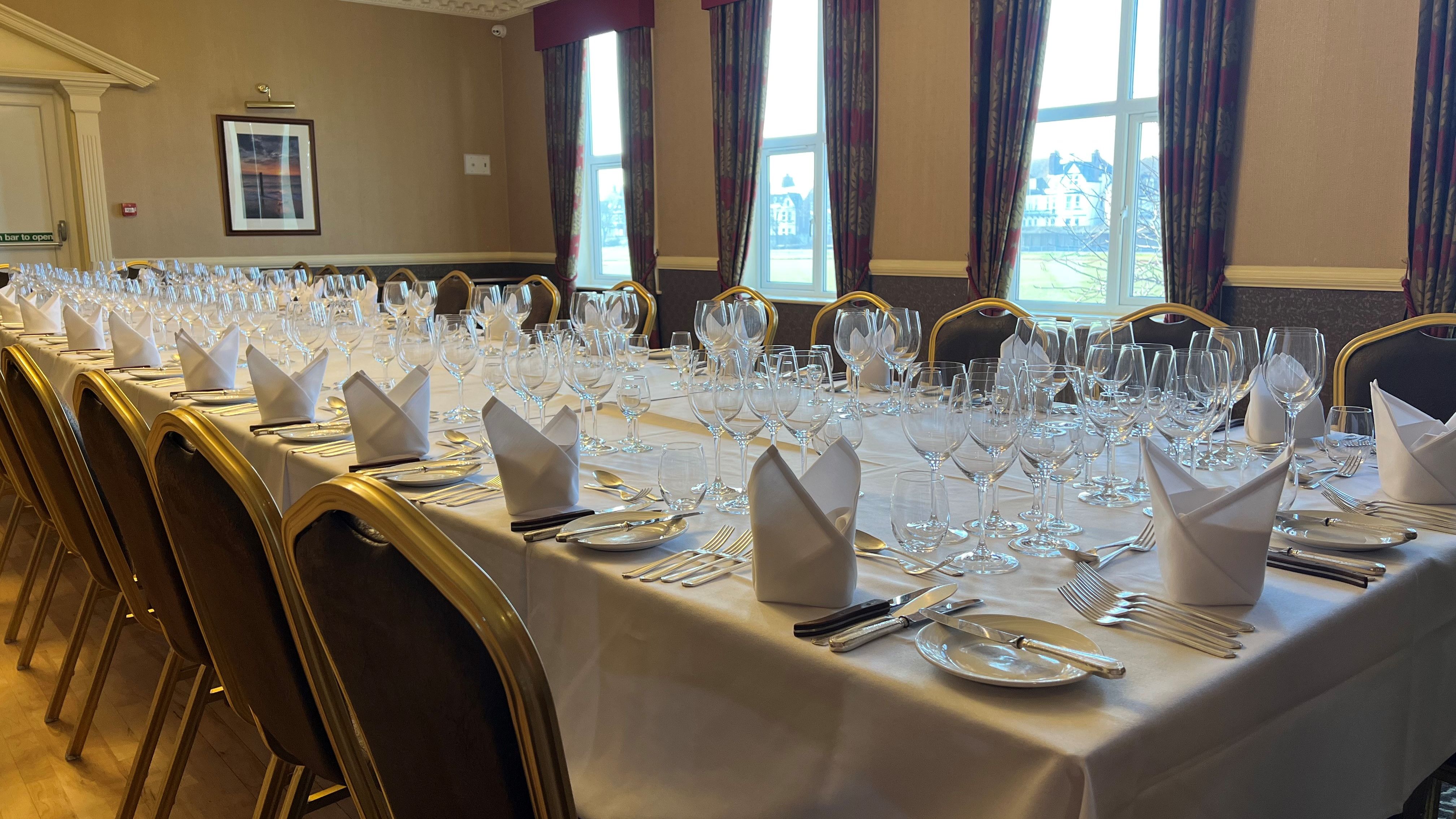 Formal dining table set with white tablecloth, multiple glasses, plates, cutlery, and folded napkins in a banquet room.