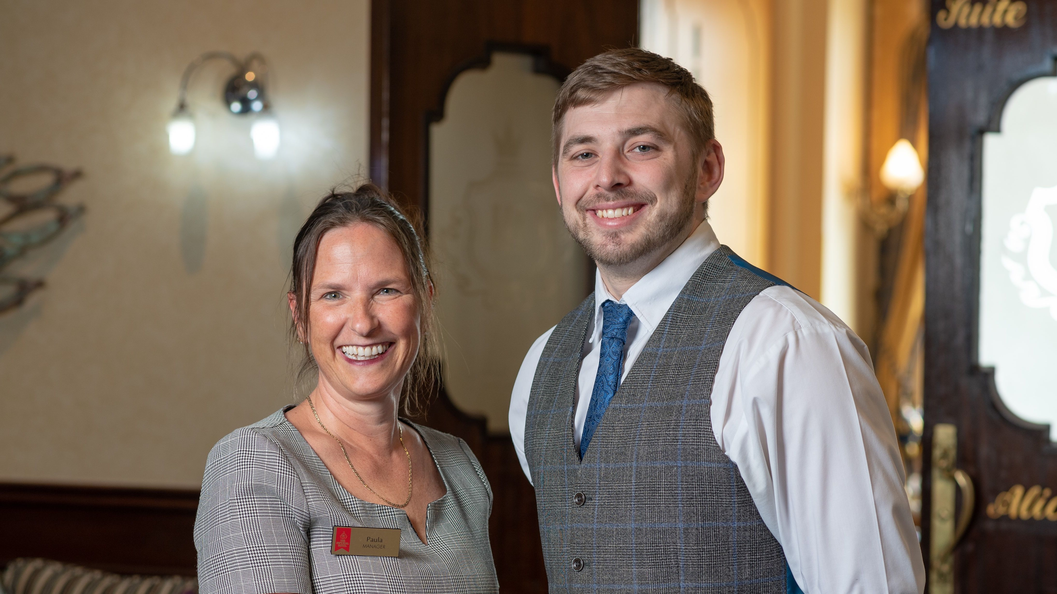 Two people standing and smiling indoors, one wearing a name tag and the other in a vest and tie.