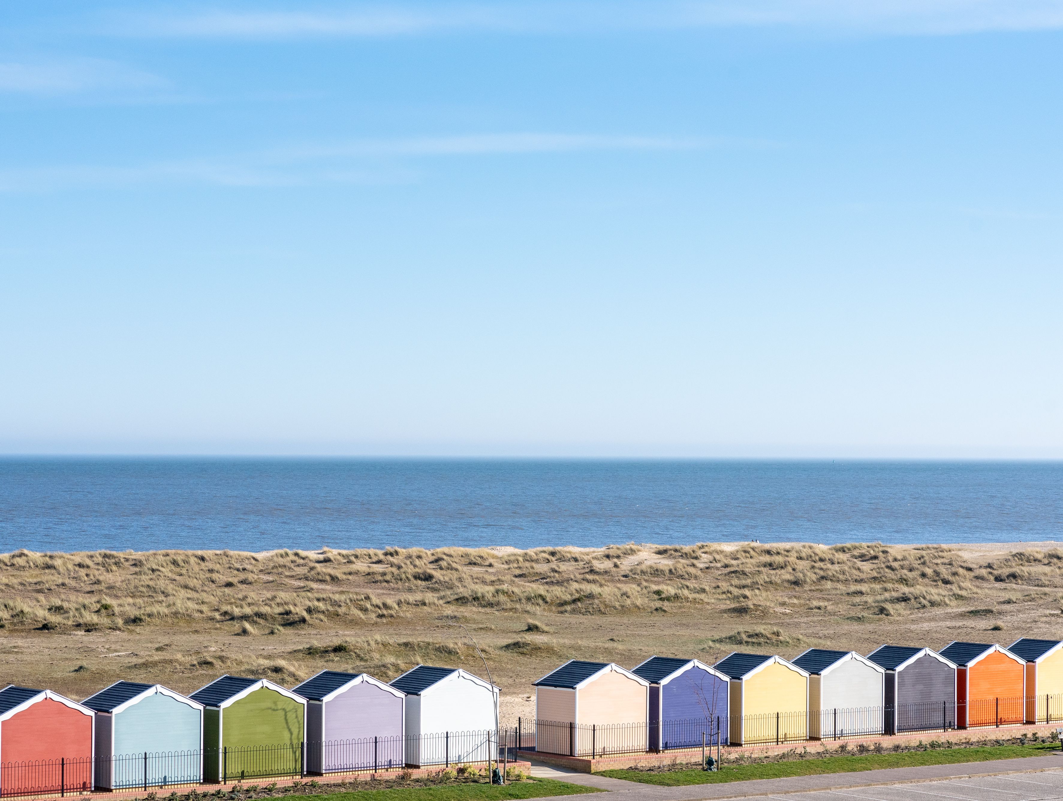 Row of colorful beach huts by the seaside with sand dunes and calm blue sea under a clear sky