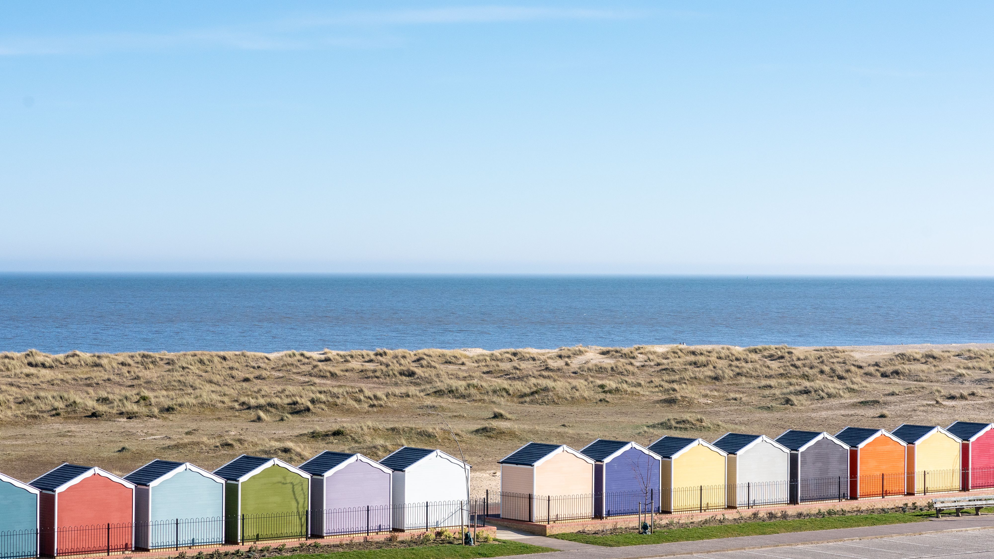 Row of colorful beach huts by the seaside with sand dunes and calm blue sea under a clear sky
