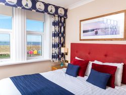 A hotel bedroom with a red headboard, blue and red pillows, nautical-themed curtains, and a sea view through the window.