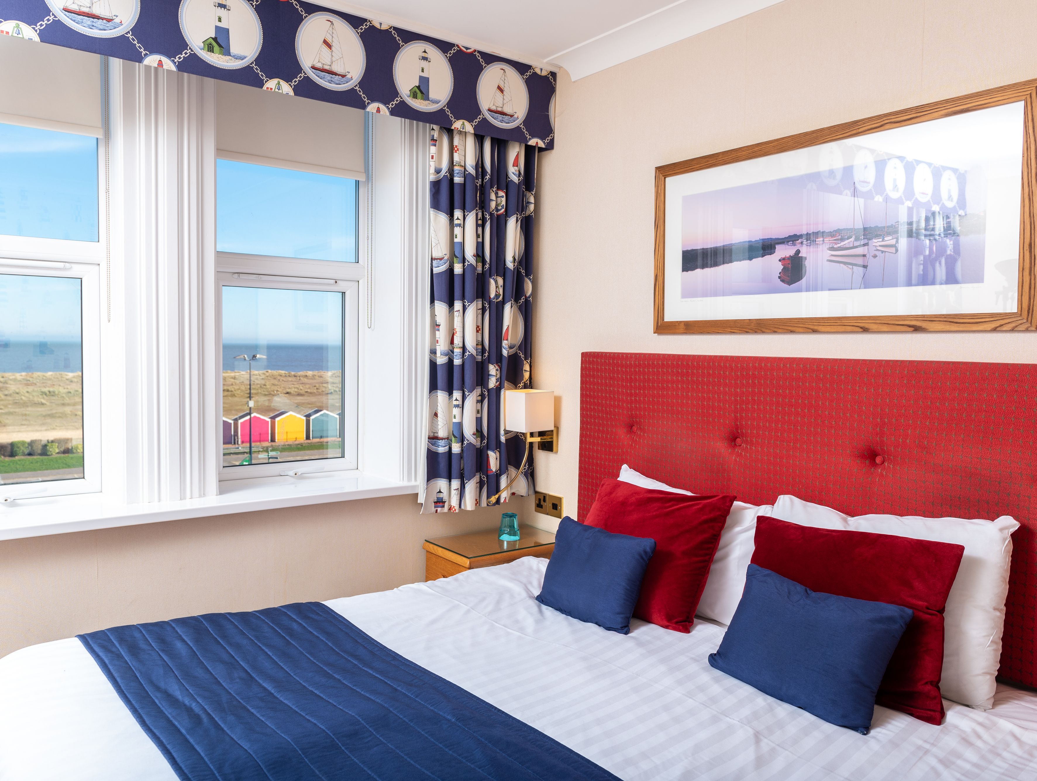 A hotel bedroom with a red headboard, blue and red pillows, nautical-themed curtains, and a sea view through the window.