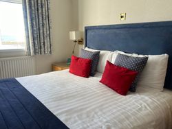 A well-made bed with a blue headboard, red and blue pillows, and a window with nautical-themed curtains.