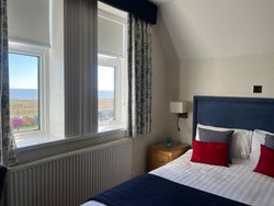 Bright hotel room with a bed, blue headboard, two windows with a sea view and colorful beach huts outside.