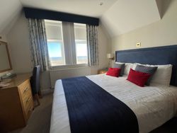 A cozy hotel room with a large bed, blue and red pillows, a desk, and windows with a sea view.
