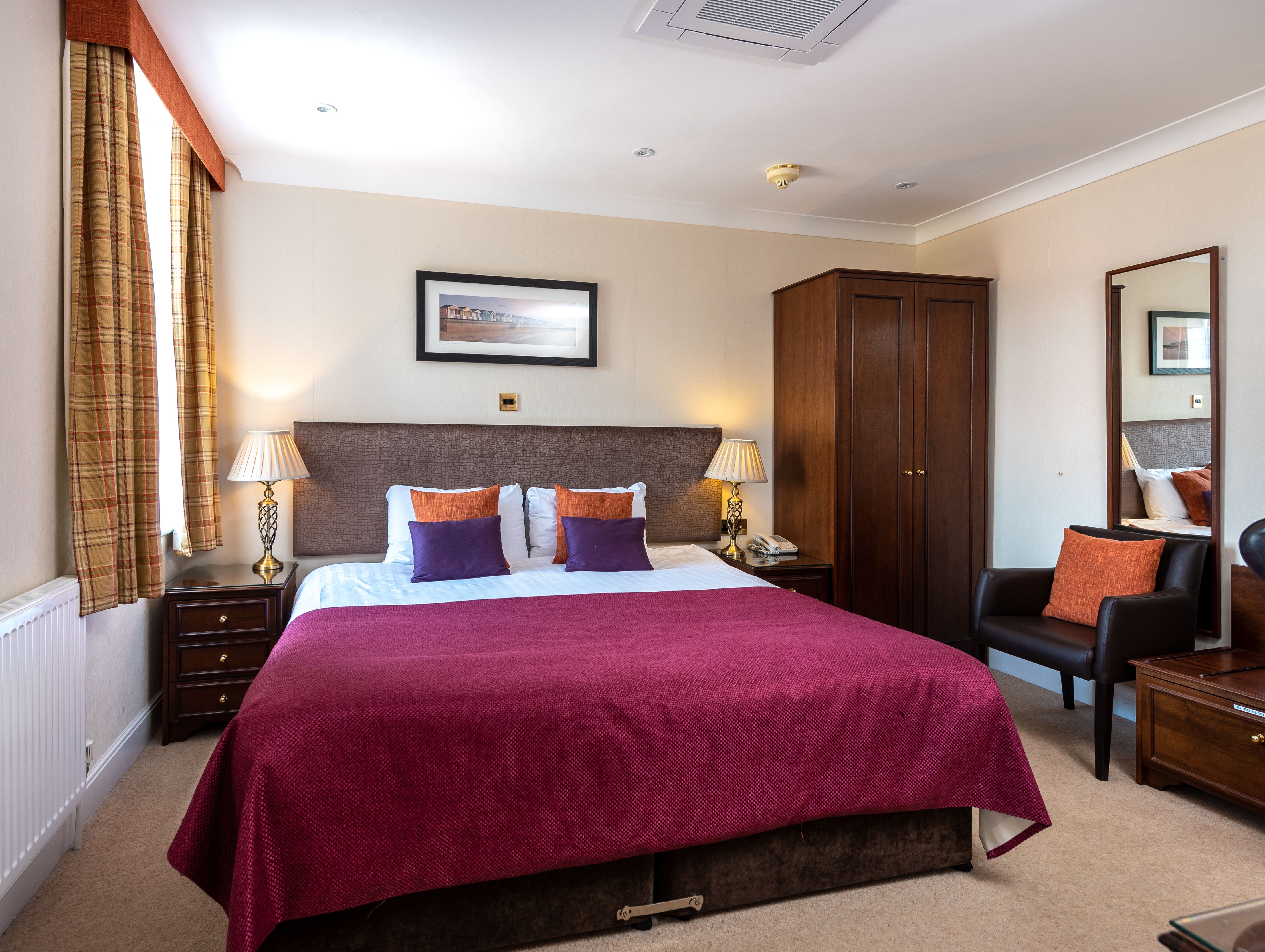 Hotel room with double bed, burgundy bedspread, wooden furniture, and window with plaid curtains