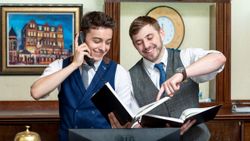 Two hotel receptionists in vests smiling while looking at a booking register, with one speaking on the phone.