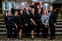 A large group of formally dressed people posing together indoors, some in uniforms and some in evening attire, on a patterned carpeted staircase.