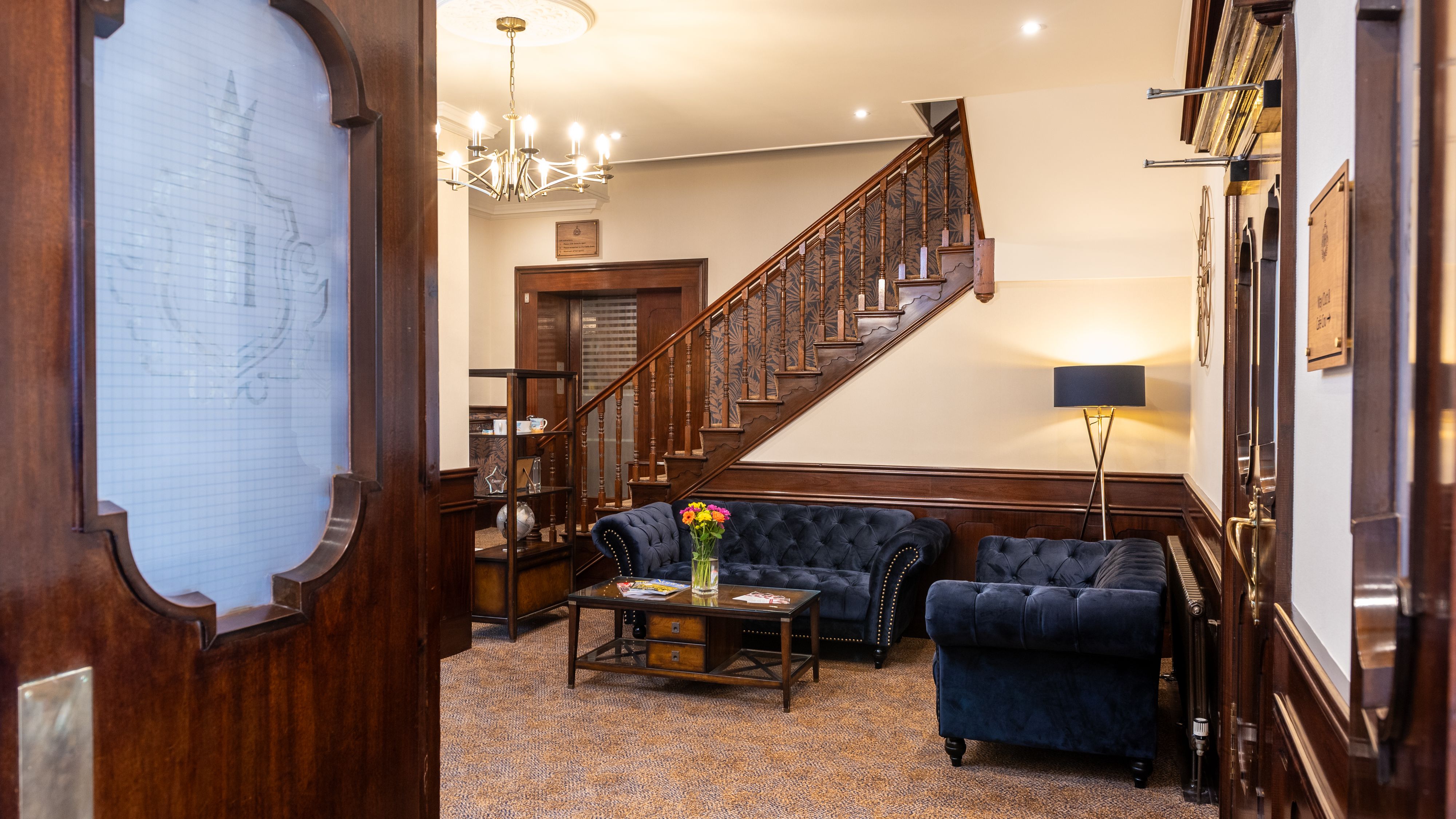 Elegant hotel lobby with dark blue sofas, wooden staircase, glass coffee table with flowers, and chandelier lighting.
