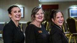 Three women standing in a row, smiling, in a conference or meeting room.
