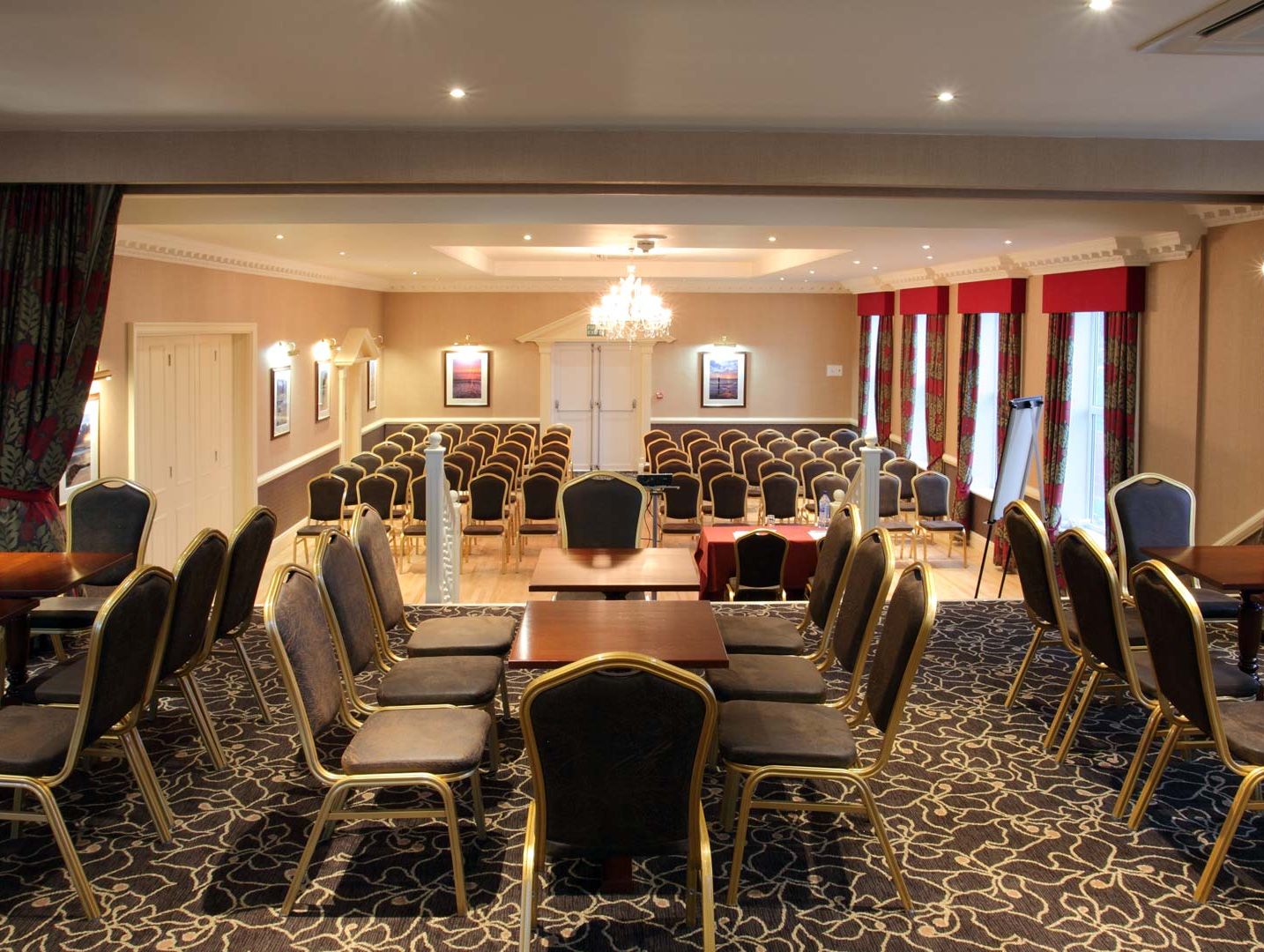 Conference room set up with rows of chairs and tables, featuring a chandelier and large windows with curtains.