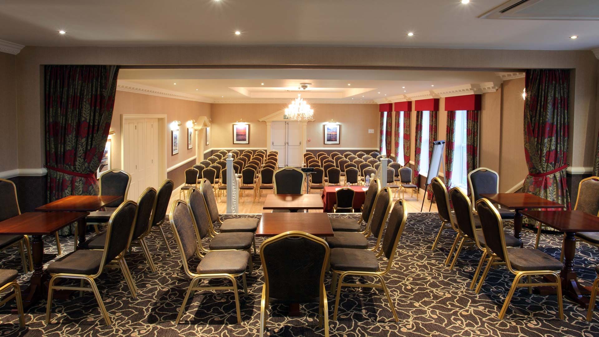 Conference room set up with rows of chairs and tables, featuring a chandelier and large windows with curtains.