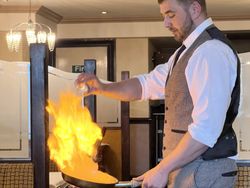 Man in vest and tie flambéing food in a pan with visible flames