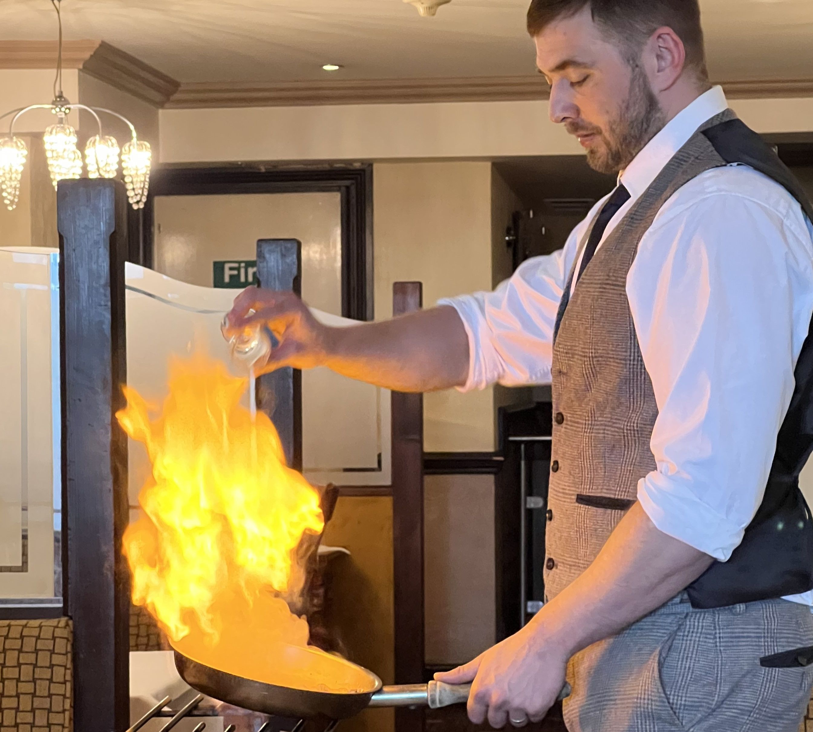 Man in vest and tie flambéing food in a pan with visible flames