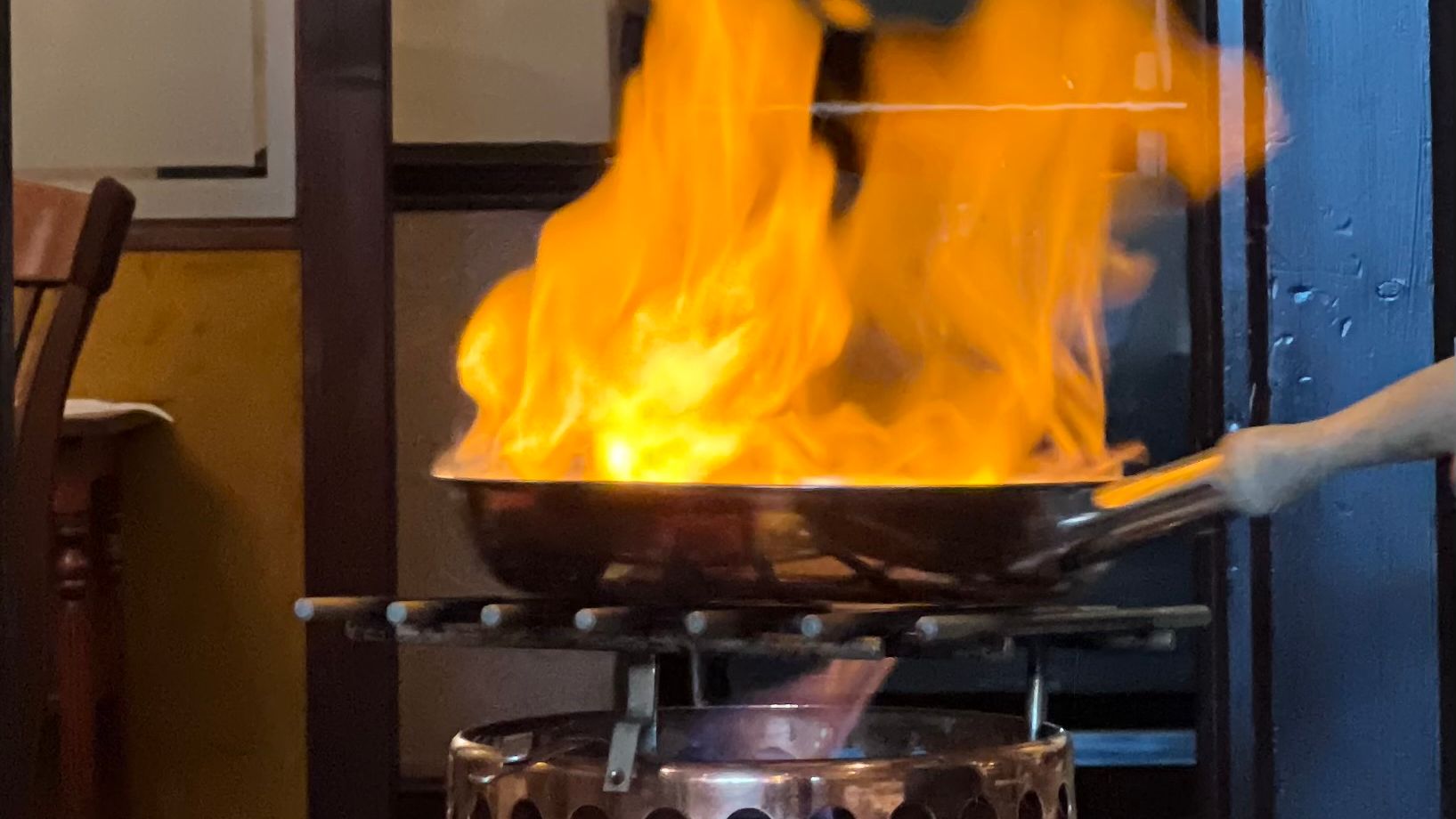 Flambé dish being prepared in a restaurant with large flames rising from a metal pan.