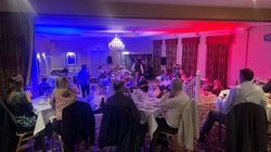 People seated at tables in a banquet hall with red and blue lighting, watching a live performance or speech.