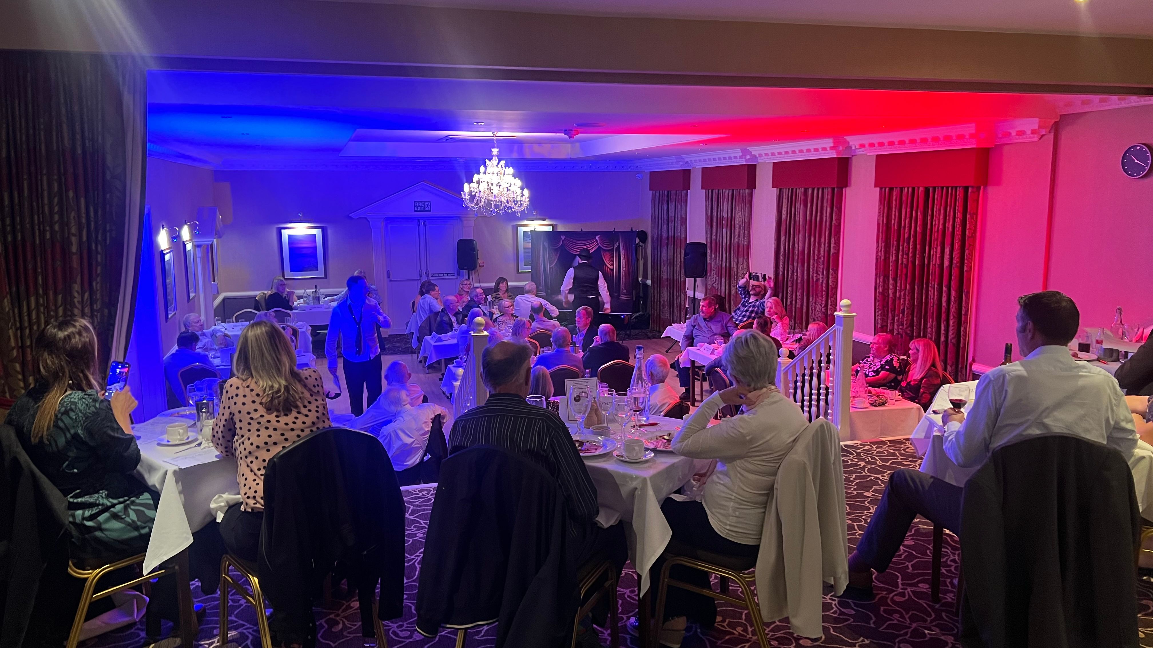 People seated at tables in a banquet hall with red and blue lighting, watching a live performance or speech.