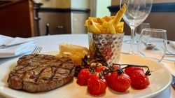 Grilled steak served with fries, roasted tomatoes, and onion rings on a white plate in a restaurant setting.