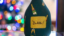 Decorative object resembling a champagne bottle with the word 'Bubbles!' on a gold label, with colorful blurred lights in the background.
