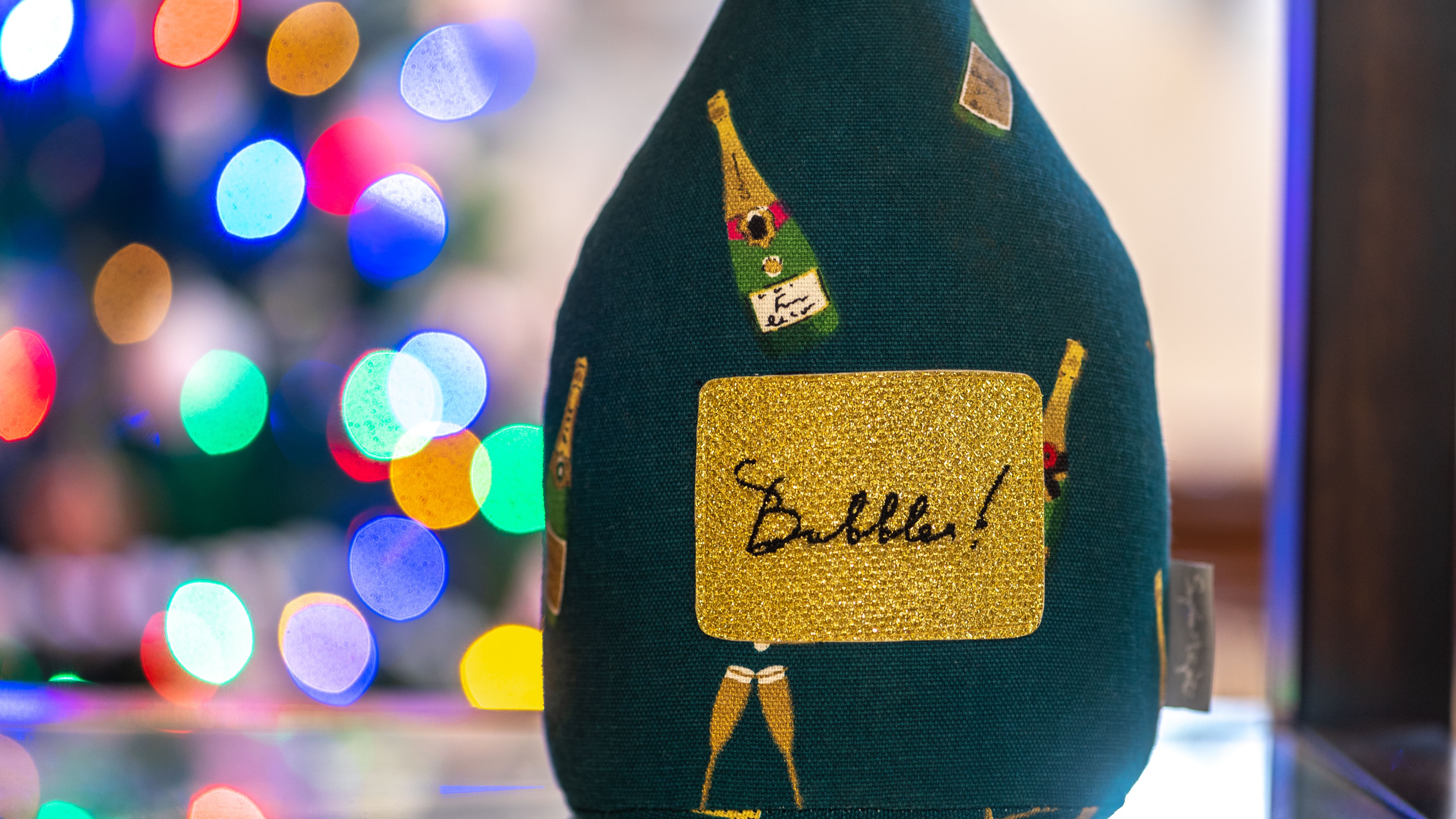 Decorative object resembling a champagne bottle with the word 'Bubbles!' on a gold label, with colorful blurred lights in the background.