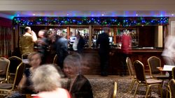 People gathered at a bar decorated with colorful string lights, with several empty chairs and tables in the foreground.