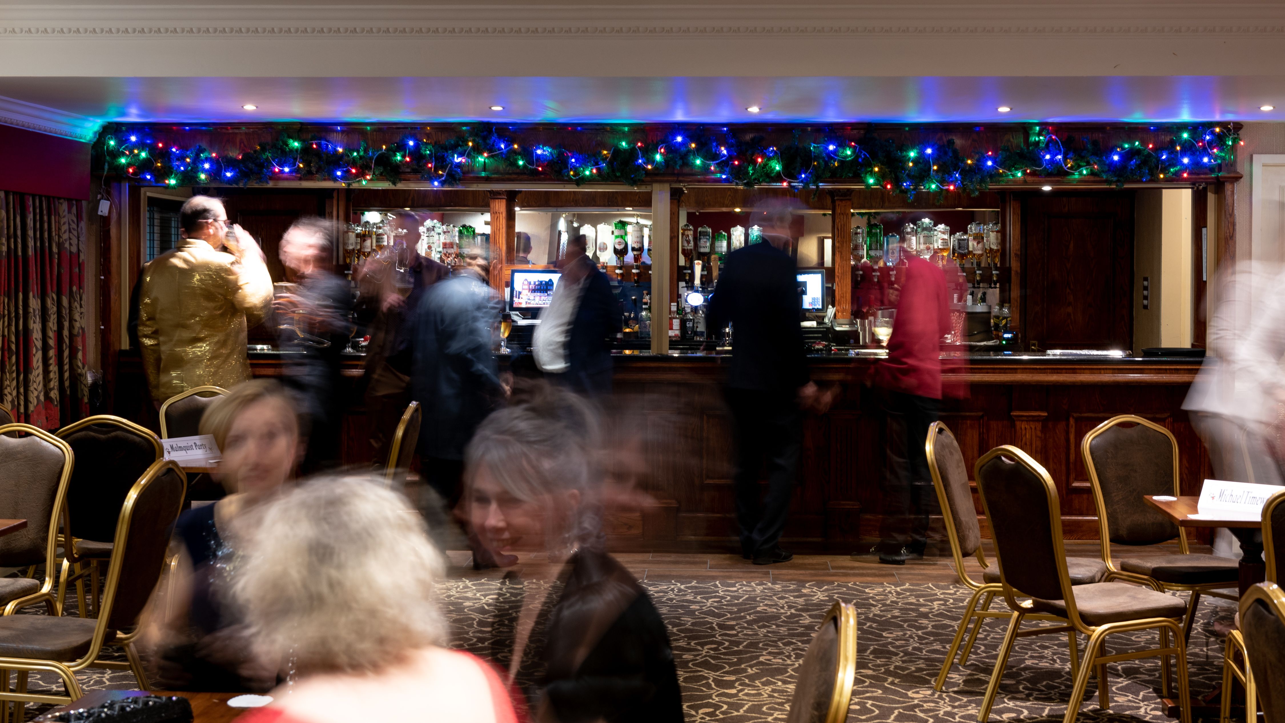 People gathered at a bar decorated with colorful string lights, with several empty chairs and tables in the foreground.