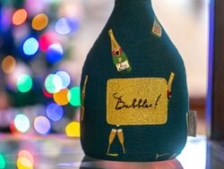 A close-up of a green fabric item decorated with champagne bottles and a sparkling gold patch labeled 'Bubbles!' with colorful bokeh lights in the background.