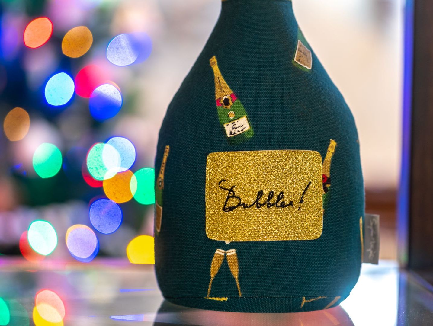A close-up of a green fabric item decorated with champagne bottles and a sparkling gold patch labeled 'Bubbles!' with colorful bokeh lights in the background.