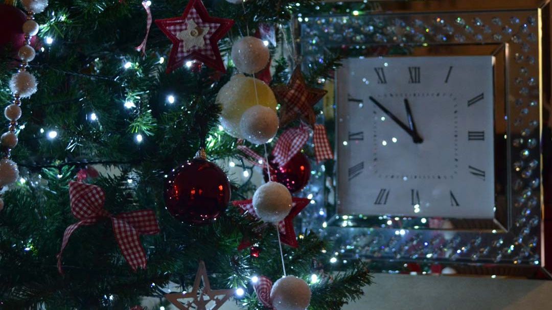 Christmas tree decorated with lights and ornaments next to a wall clock under a staircase