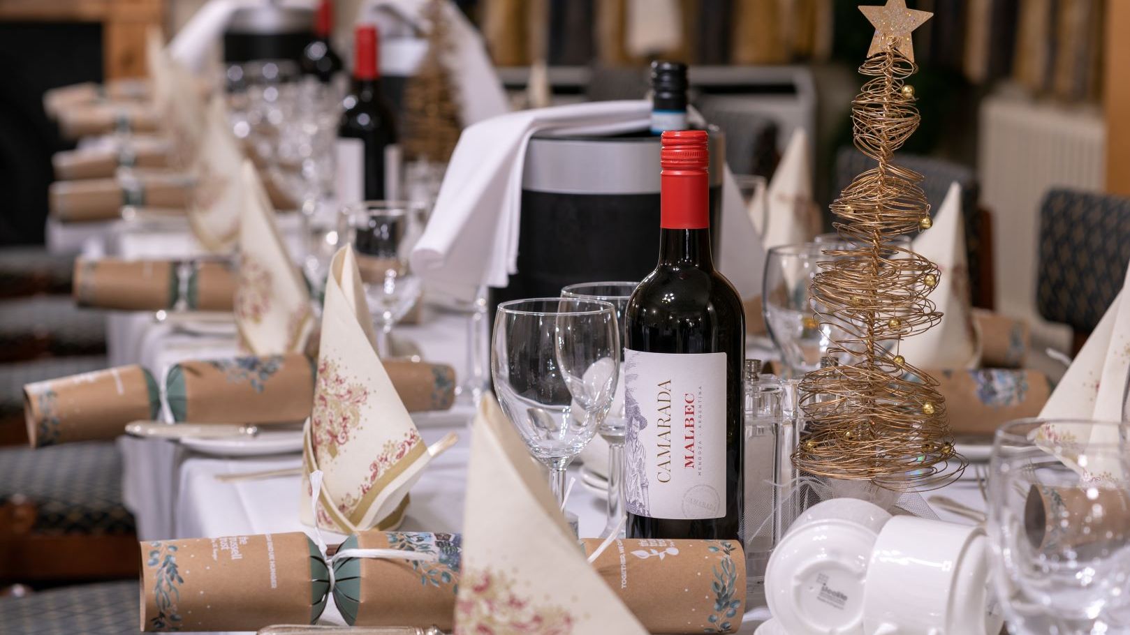 A festive dining table set with wine, glasses, Christmas crackers, napkins, and gold wire Christmas tree decorations.