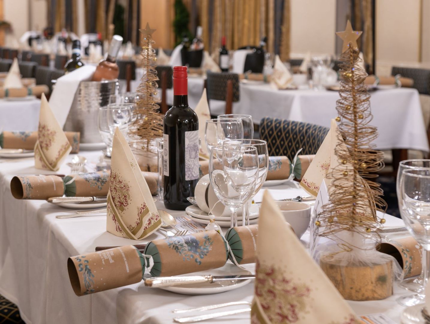 Festive dining table set with wine, glasses, napkins, Christmas crackers and gold wire tree decorations.