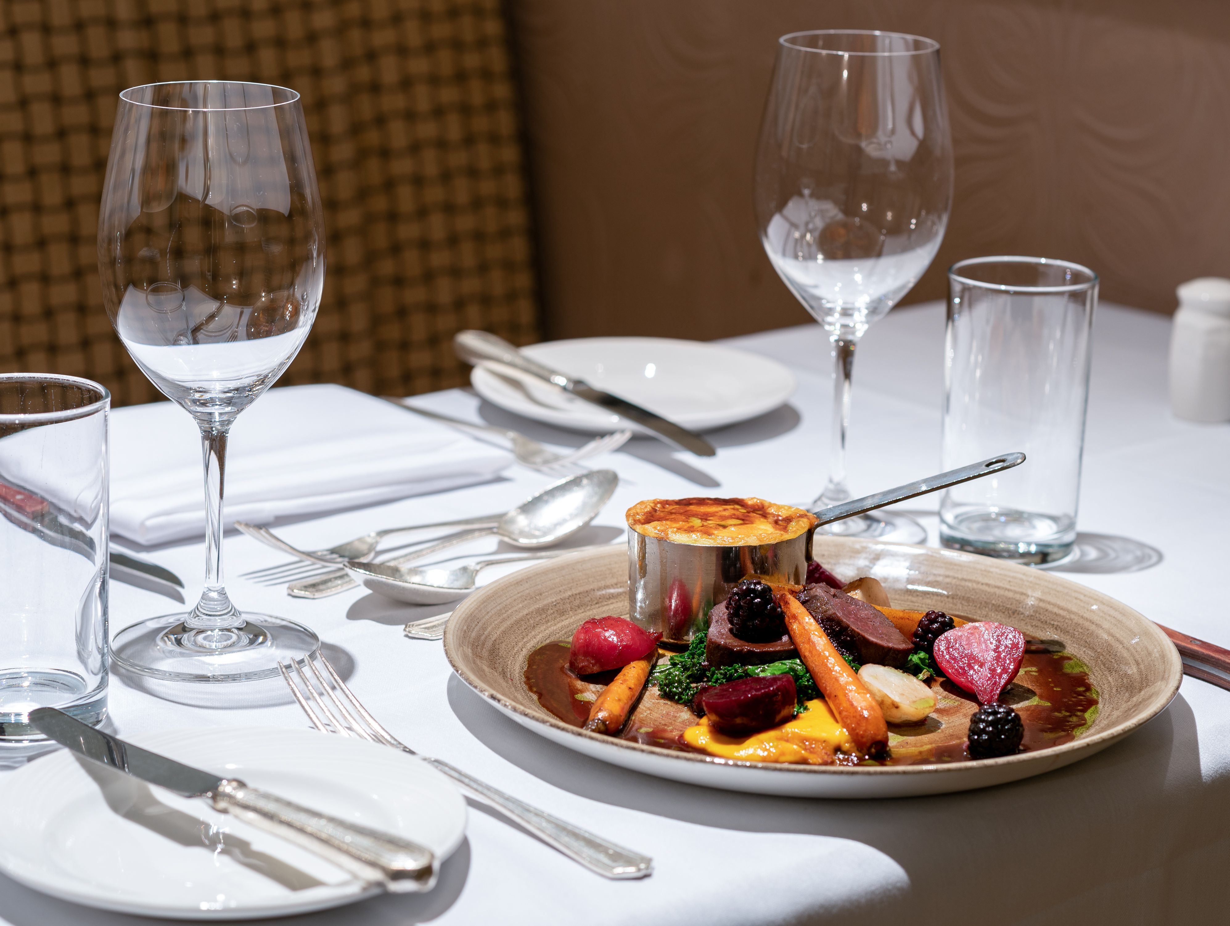 Elegant restaurant table setting with gourmet plated dish featuring vegetables, sauce, and a baked side, along with wine glasses and cutlery.