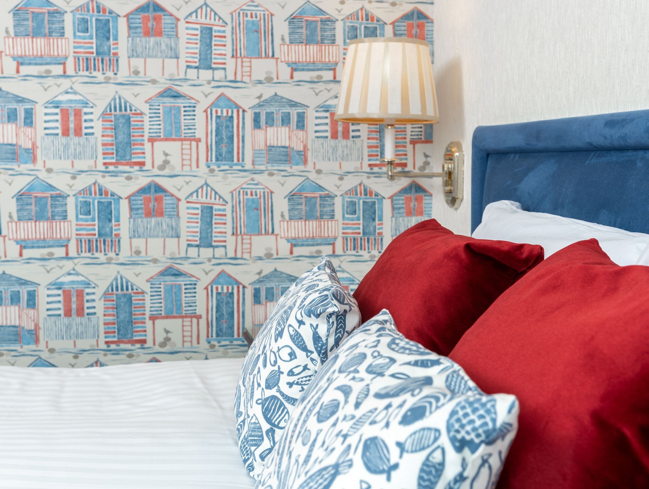 Close-up of a bed with red and blue pillows, in front of beach hut themed wallpaper and a wall lamp
