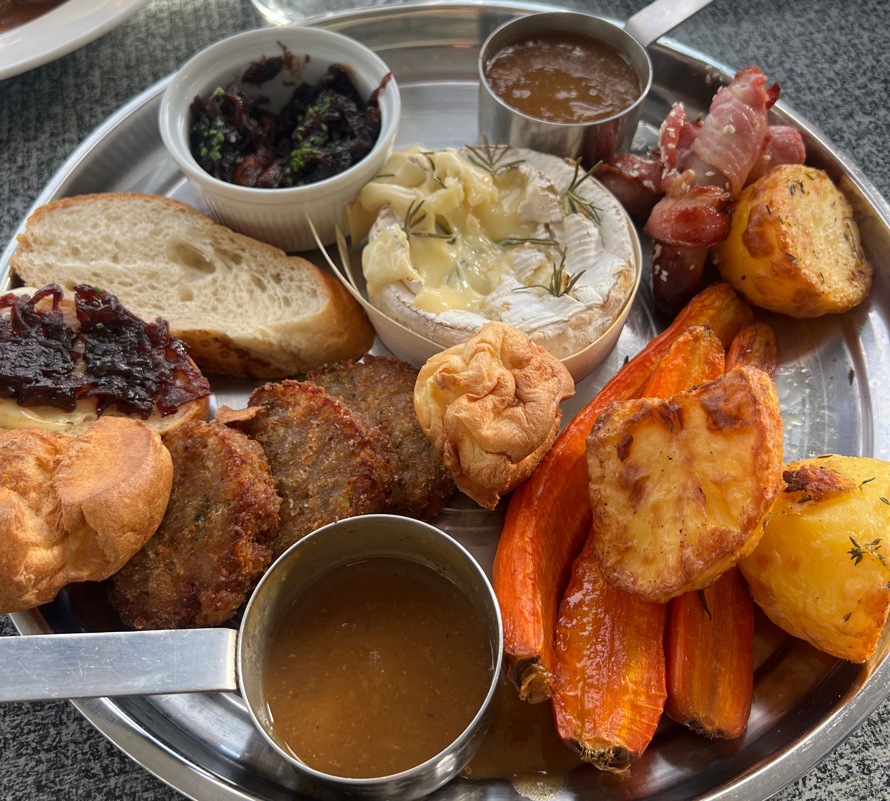 A festive sharing platter with roasted potatoes, carrots, Yorkshire puddings, baked Camembert, bread, stuffing, bacon-wrapped sausages, assorted sauces, and onion chutney.