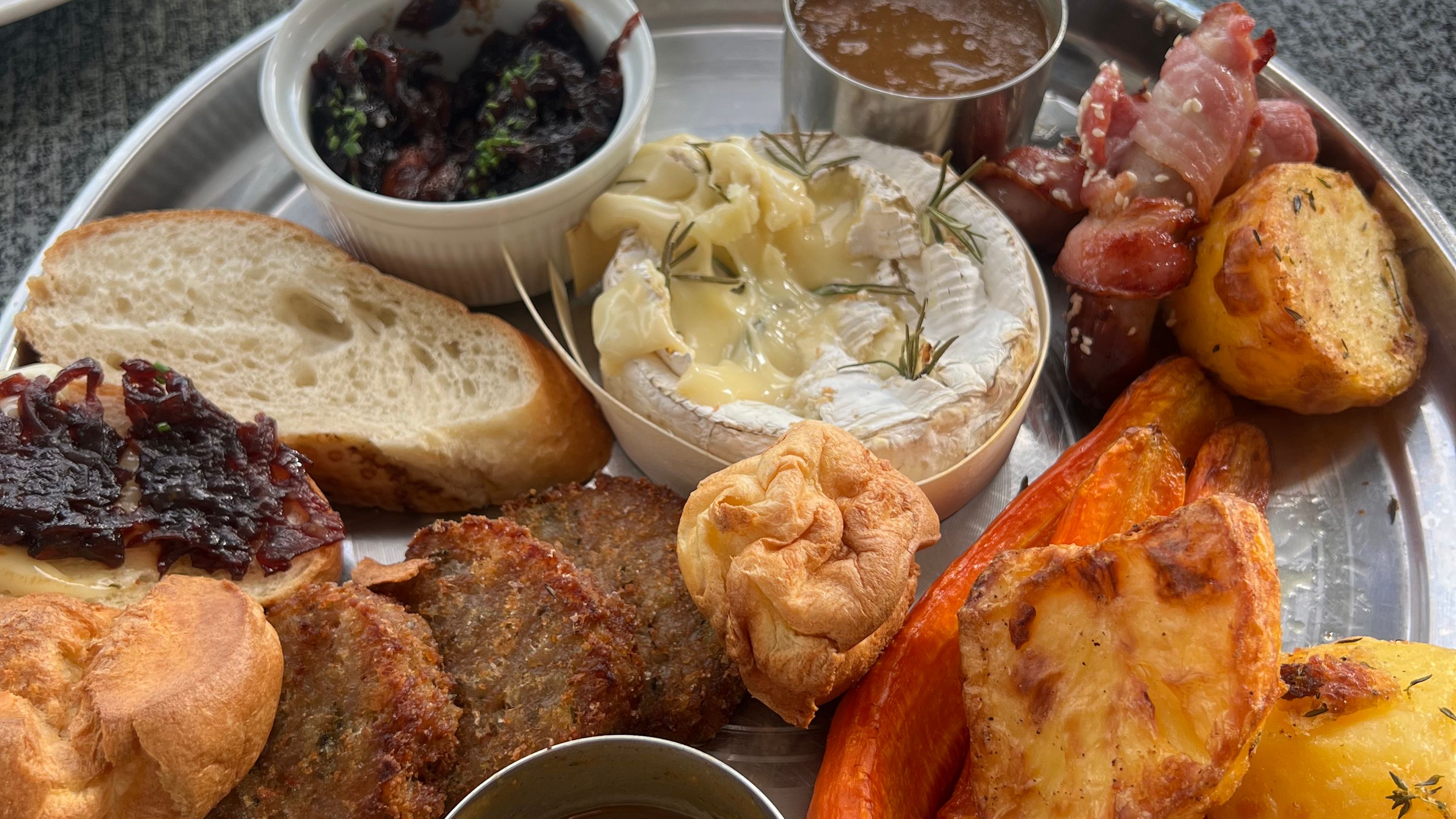 A festive sharing platter with roasted potatoes, carrots, Yorkshire puddings, baked Camembert, bread, stuffing, bacon-wrapped sausages, assorted sauces, and onion chutney.