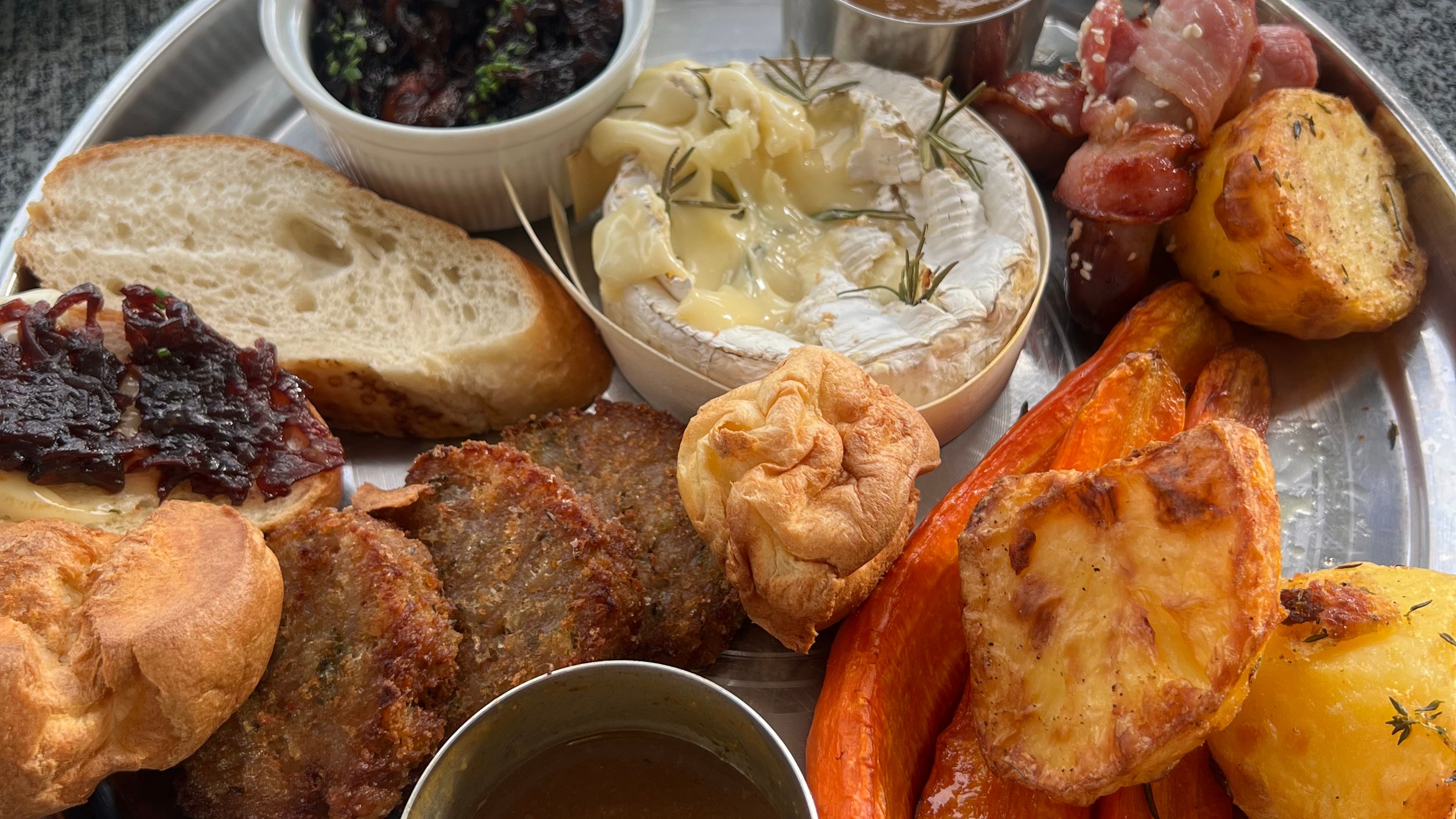 A festive sharing platter with roasted potatoes, carrots, Yorkshire puddings, baked Camembert, bread, stuffing, bacon-wrapped sausages, assorted sauces, and onion chutney.