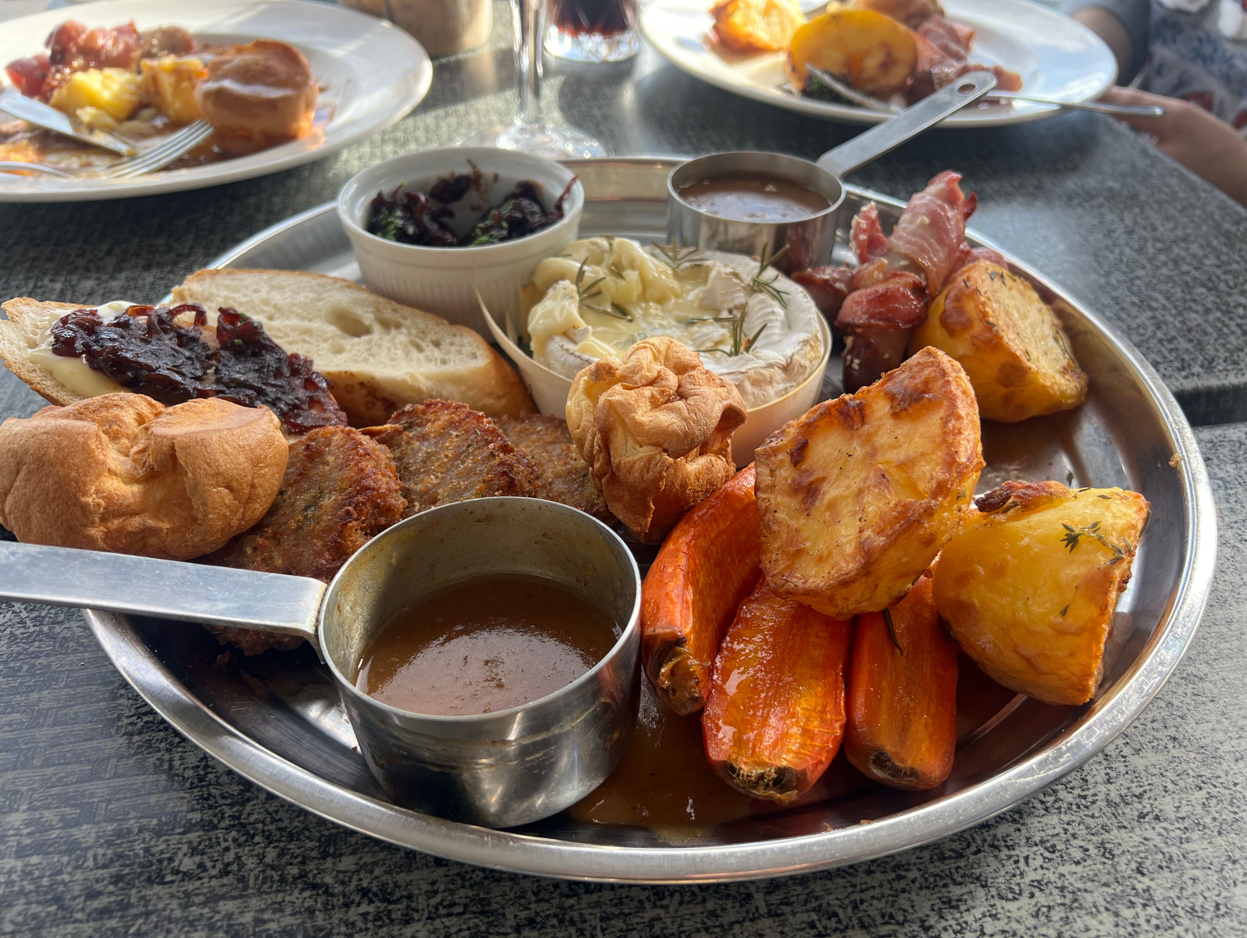 A festive sharing platter with roast potatoes, carrots, Yorkshire pudding, brie, bread, jam, bacon-wrapped items, and dips on a metal plate.