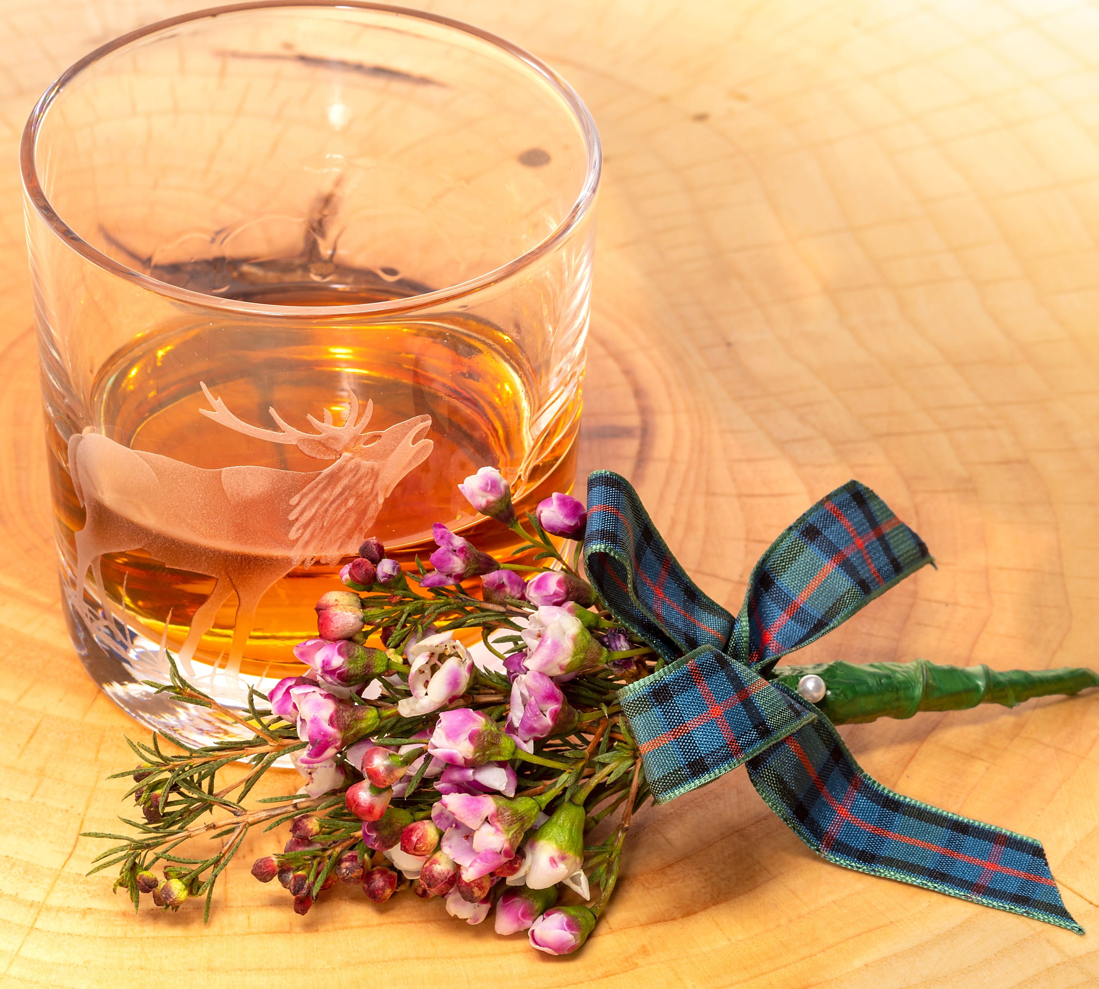 A glass of whisky with a stag engraving, next to a small bouquet of purple flowers tied with a tartan ribbon.