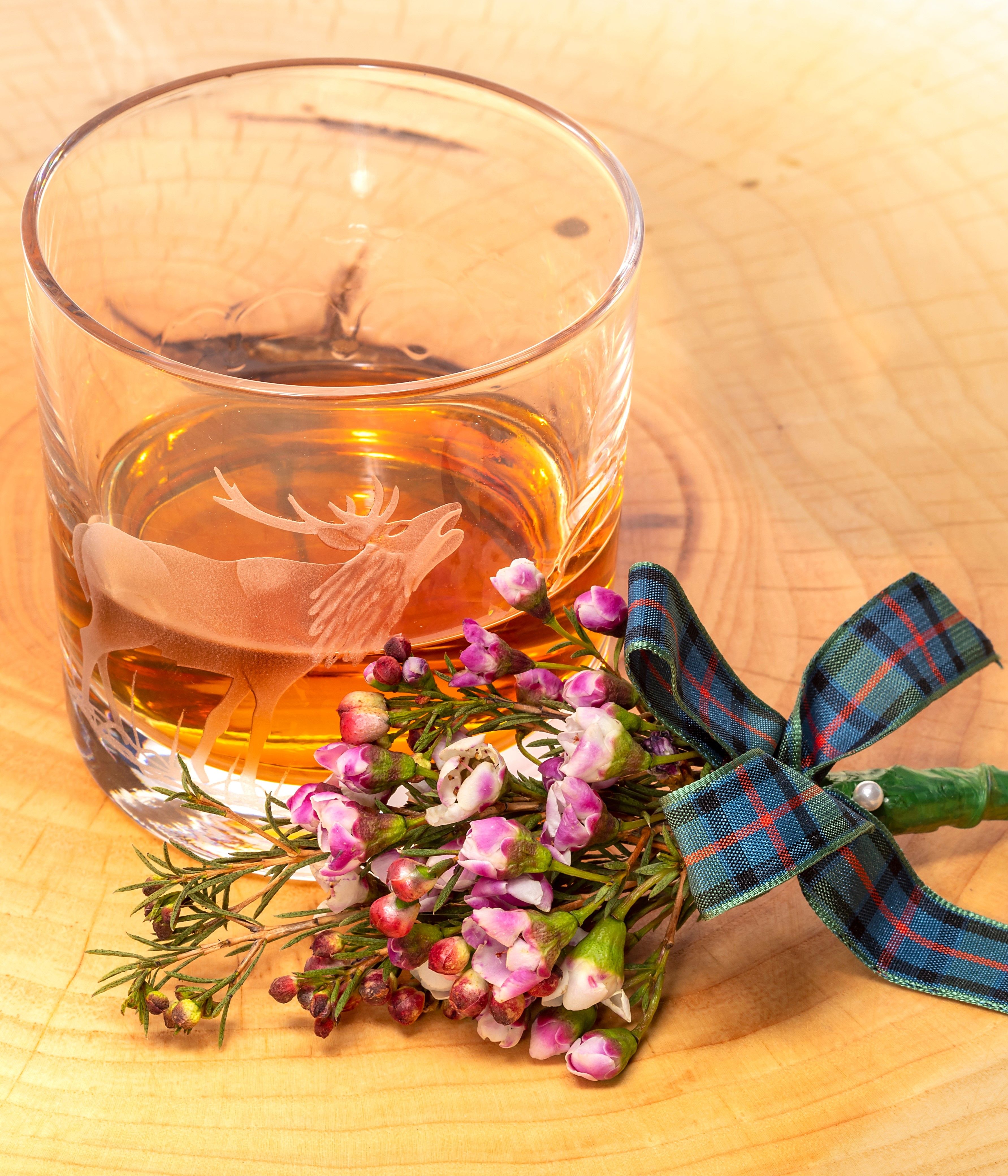 A glass of whisky with a stag engraving, next to a small bouquet of purple flowers tied with a tartan ribbon.