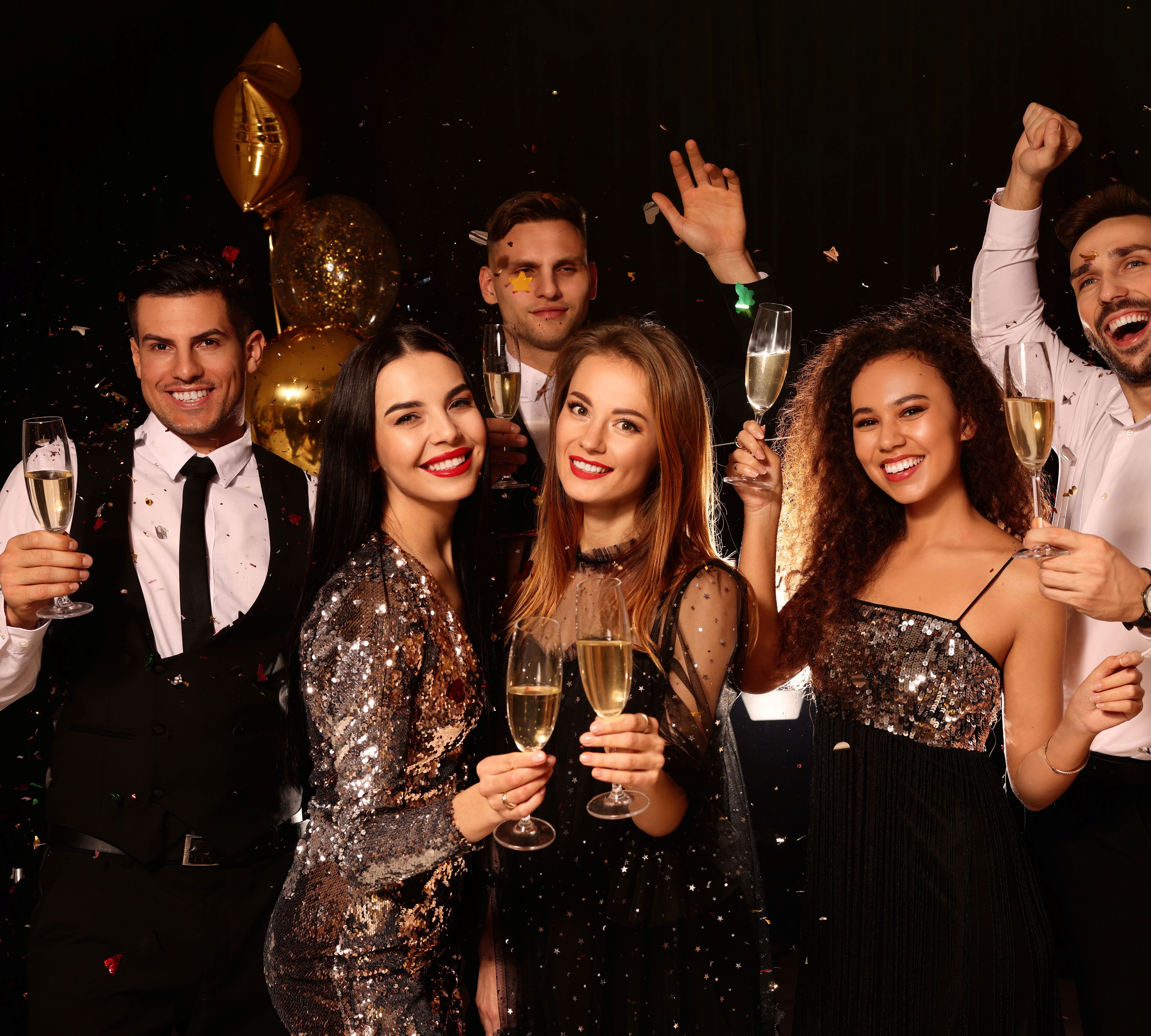 Group of happy young adults celebrating with champagne at a party, surrounded by gold balloons and confetti.