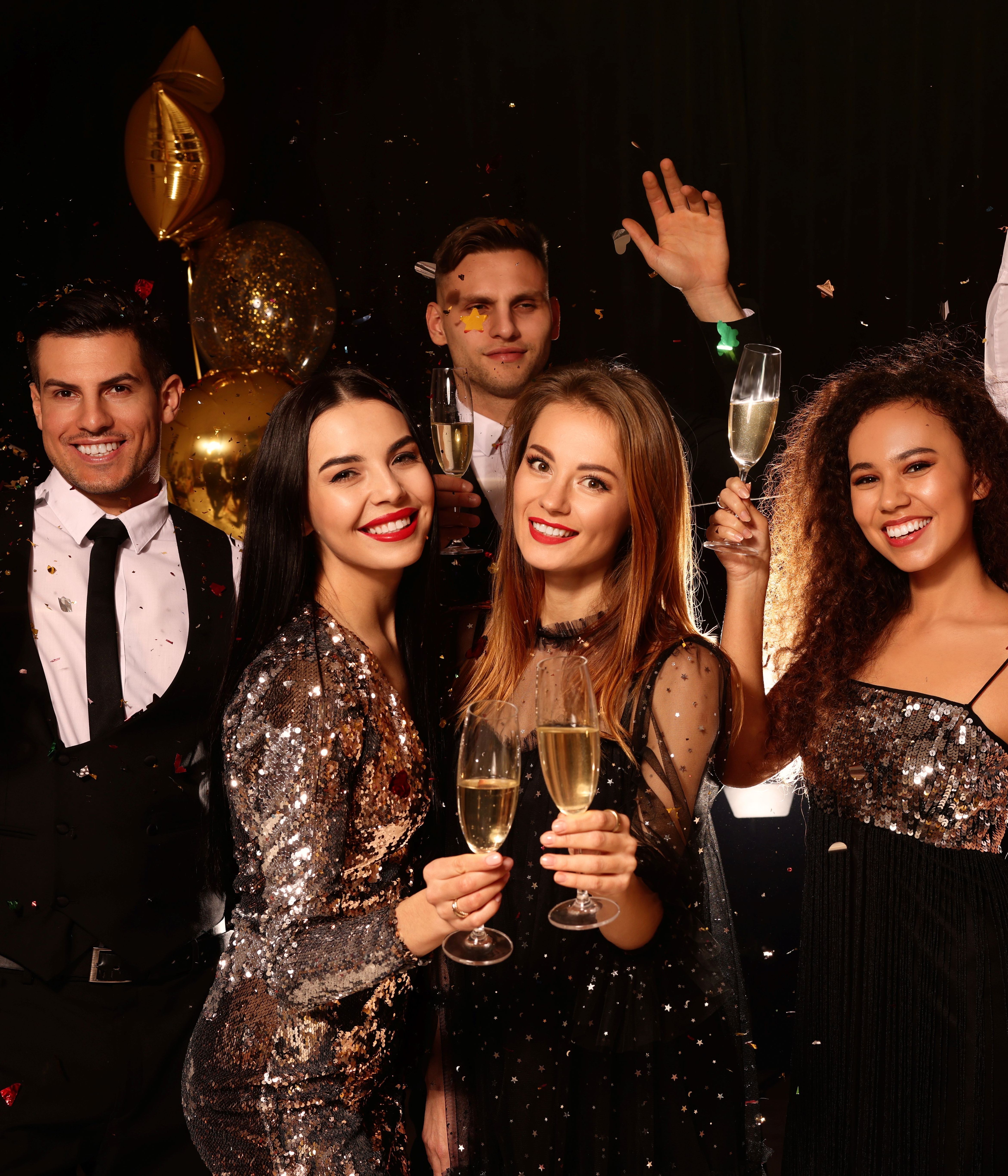 Group of happy young adults celebrating with champagne at a party, surrounded by gold balloons and confetti.
