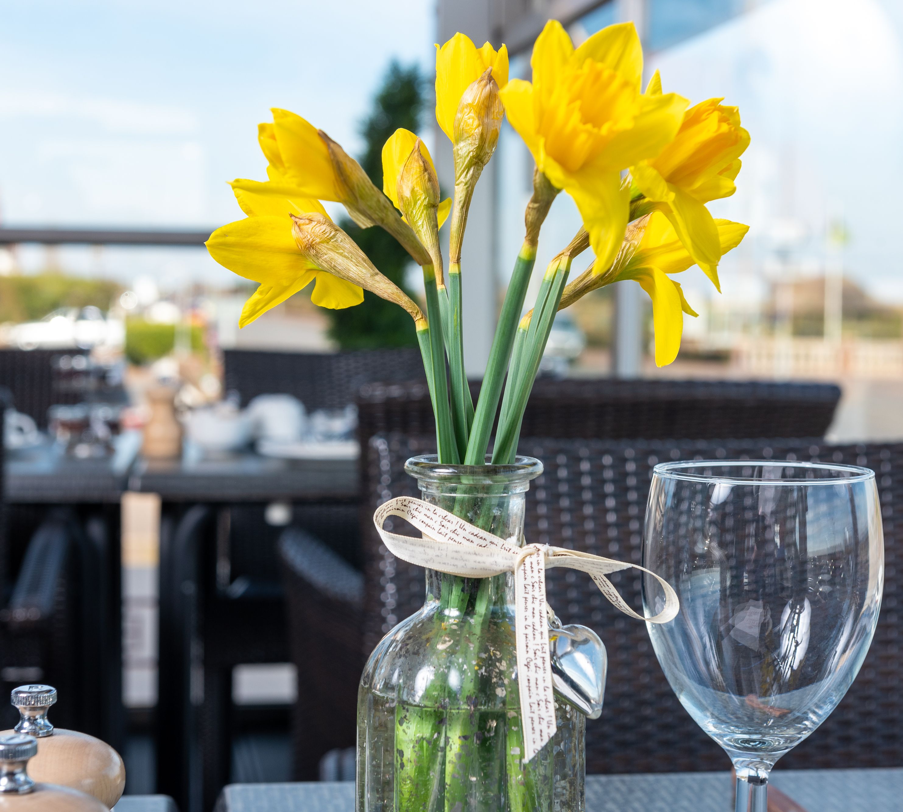 Yellow daffodils in a glass vase on an outdoor restaurant table, with a wine glass and blurred background.