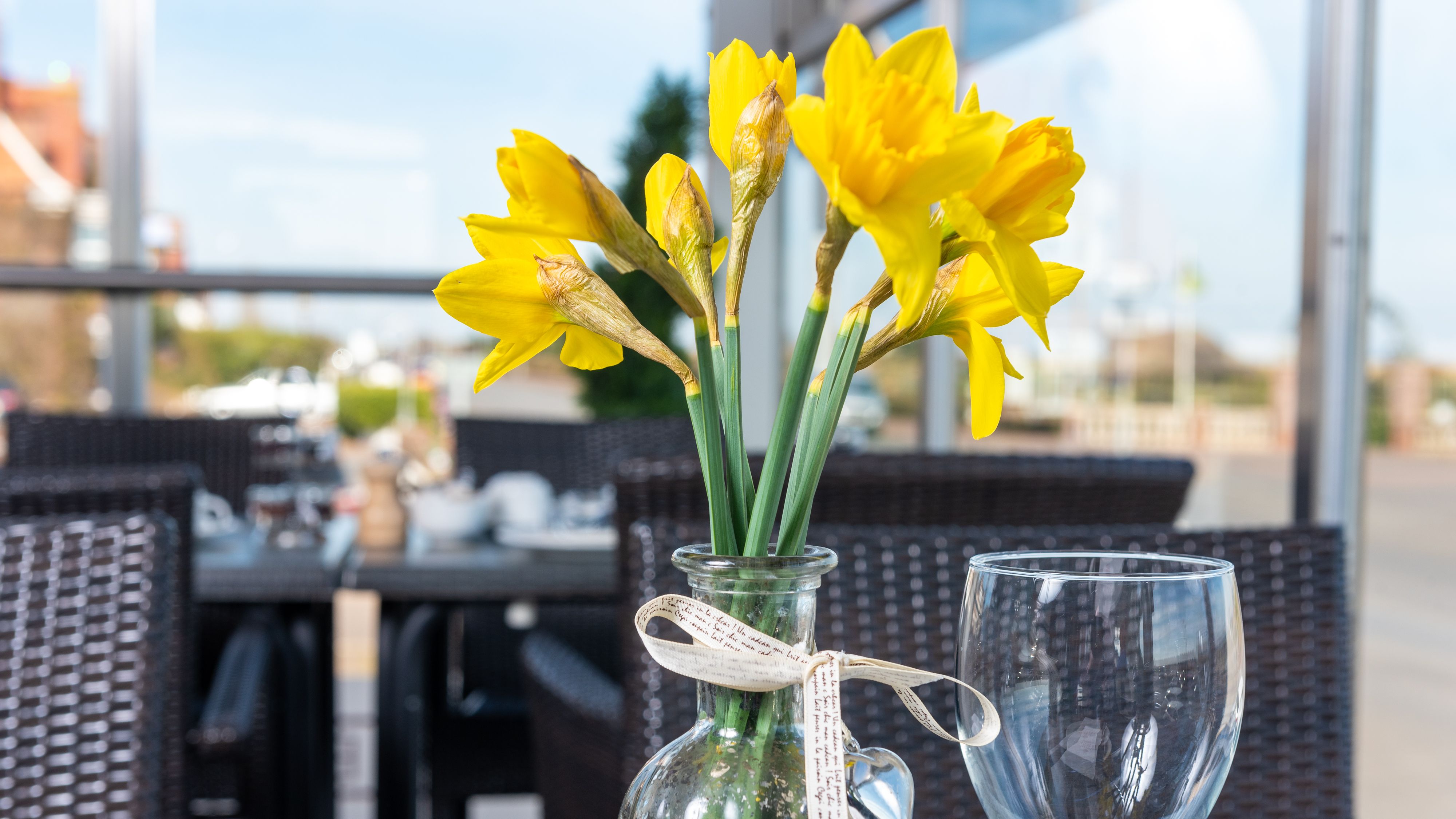 Yellow daffodils in a glass vase on an outdoor restaurant table, with a wine glass and blurred background.