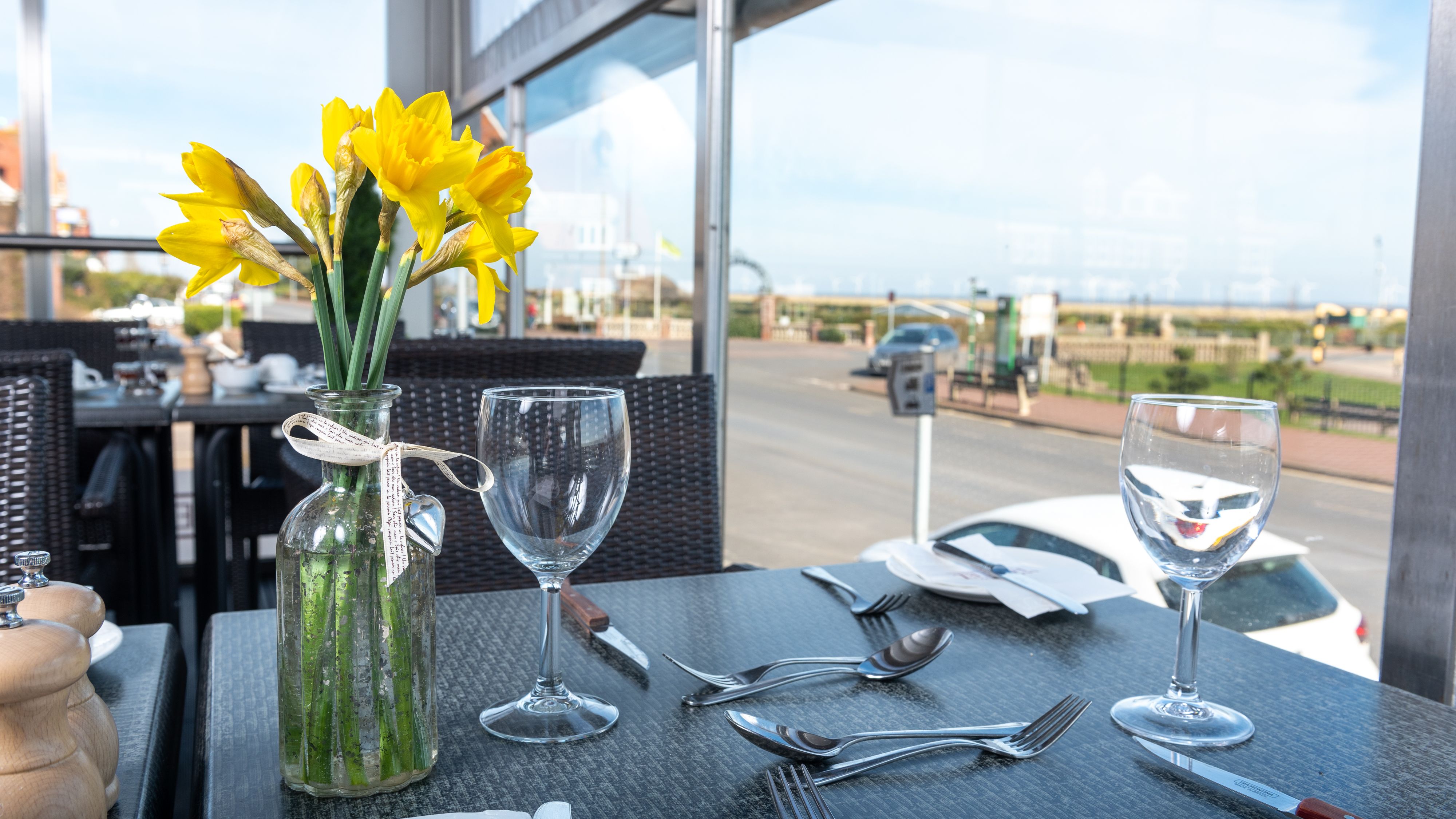 Table set for two with daffodils in a glass vase, overlooking a bright street through large windows.